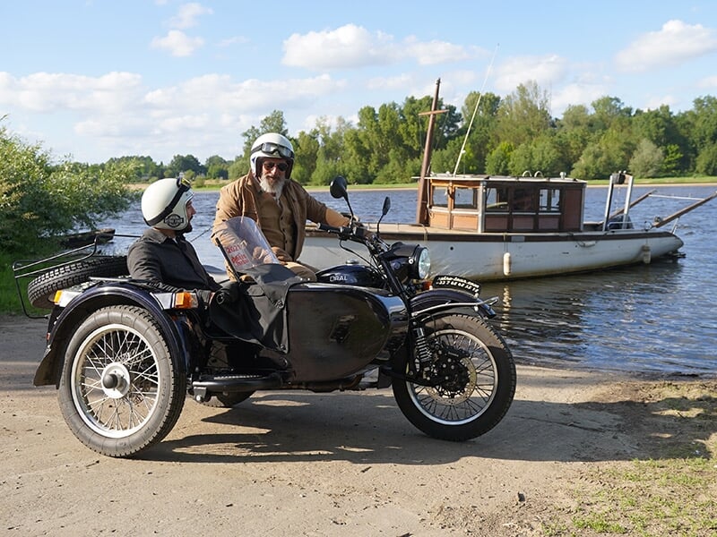 Un homme et un enfant assis dans un side-car noir, garé près d'une rivière avec un bateau en bois amarré, sous un ciel bleu avec des nuages.