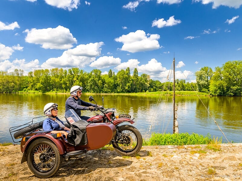 Une moto avec side-car marron stationnée près d'une rivière calme, avec un voilier traditionnel ancré en arrière-plan, sous un ciel bleu éclatant.