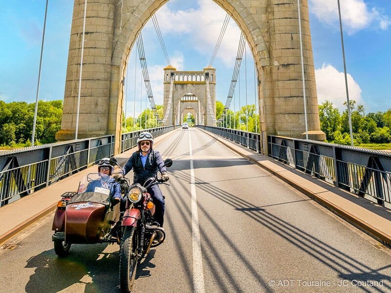  Une moto avec side-car marron traversant un grand pont suspendu, entourée d'une belle architecture de style historique sous un ciel bleu dégagé.