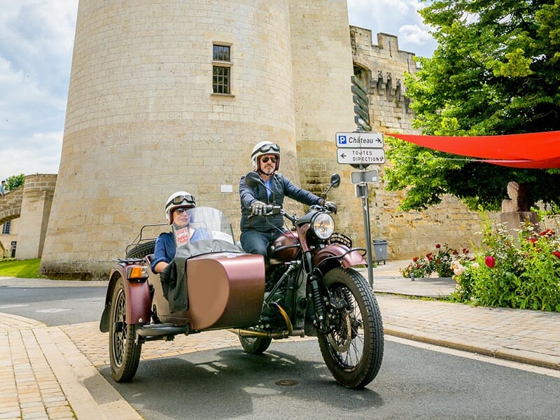 Un duo en moto avec side-car marron, roulant près d'une forteresse médiévale en pierre avec une tour imposante, lors d'une journée ensoleillée.
