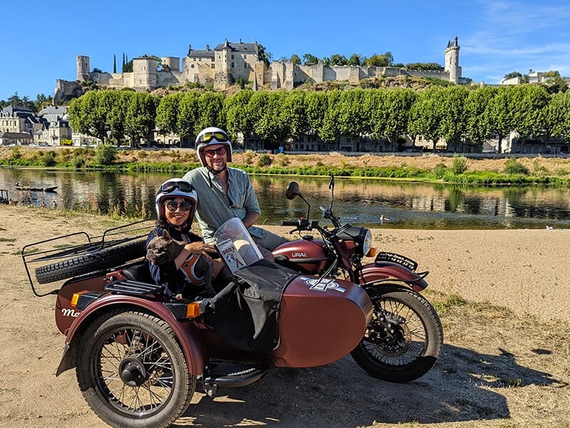  Un homme et une femme en side-car marron stationnés le long d'une rivière, avec une vue panoramique sur une ville médiévale fortifiée au sommet d'une colline.