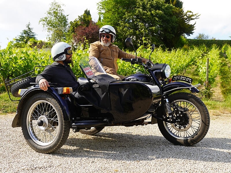  Deux personnes en side-car noir, posant dans un vignoble, sous un ciel ensoleillé avec des rangées de vignes en arrière-plan.