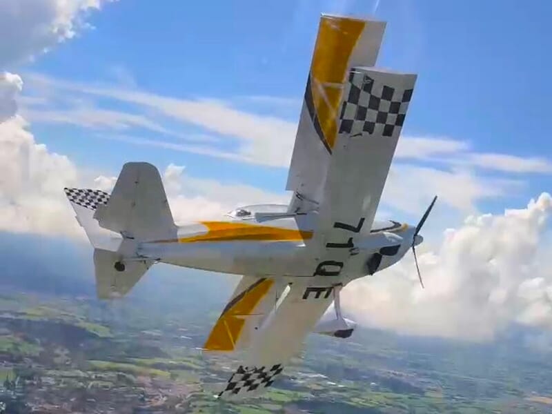 Avion biplan blanc et jaune en vol avec des motifs en damier sur l’aile arrière, évoluant dans un ciel bleu.