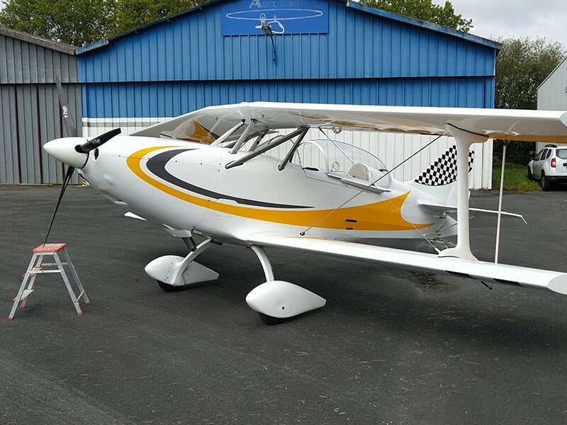 Avion biplan blanc et jaune stationné sur le tarmac devant un hangar bleu, prêt pour un vol.