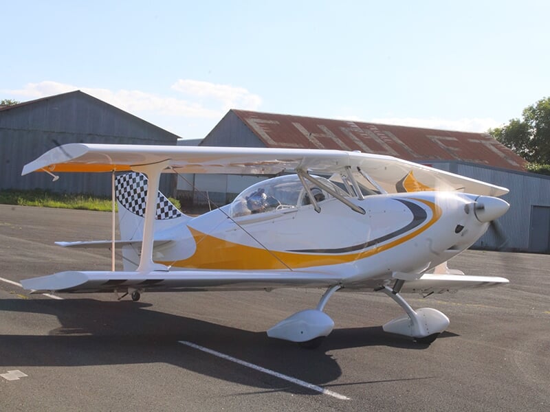 Avion biplan blanc et jaune garé sur une piste, ensoleillé, avec des bâtiments industriels en arrière-plan.