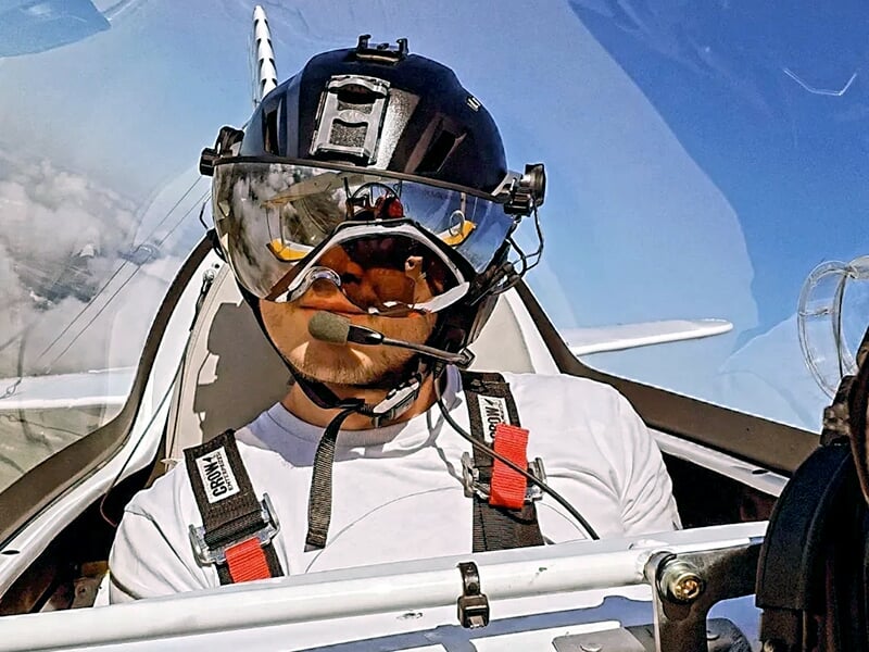 Pilote en tenue d’aviateur avec casque et visière, assis dans le cockpit, regardant droit devant lui avec un fond de ciel bleu.