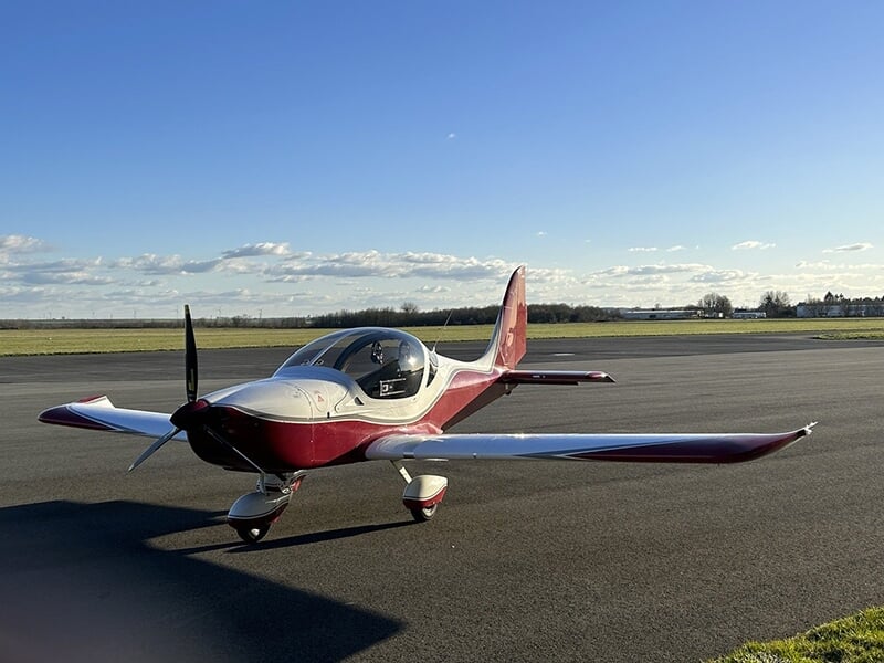 Avion léger rouge et blanc stationné sur une piste d'aviation sous un ciel bleu clair.