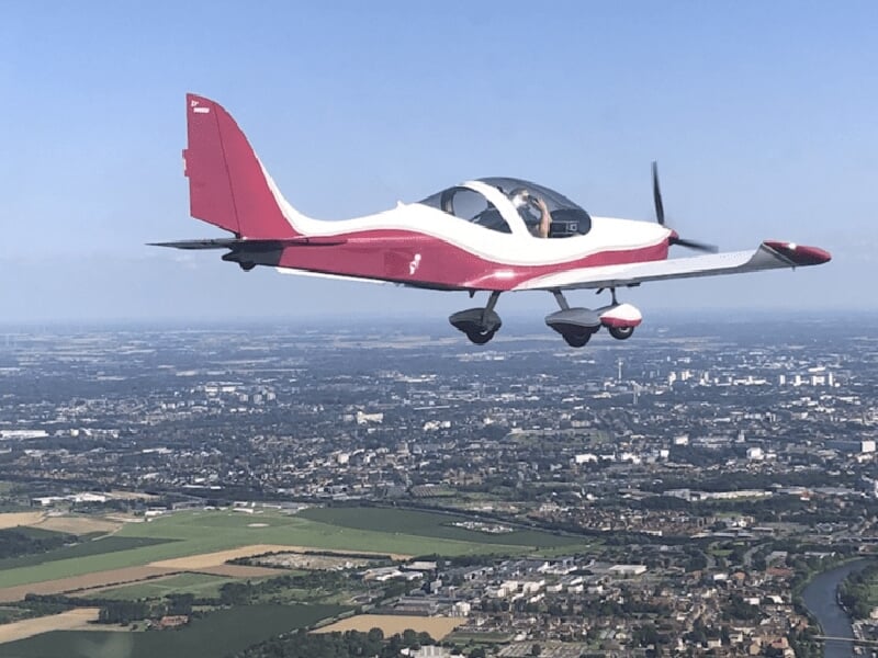 Avion léger en vol au-dessus d'une zone urbaine et rurale, avec des champs et des bâtiments visibles au sol.