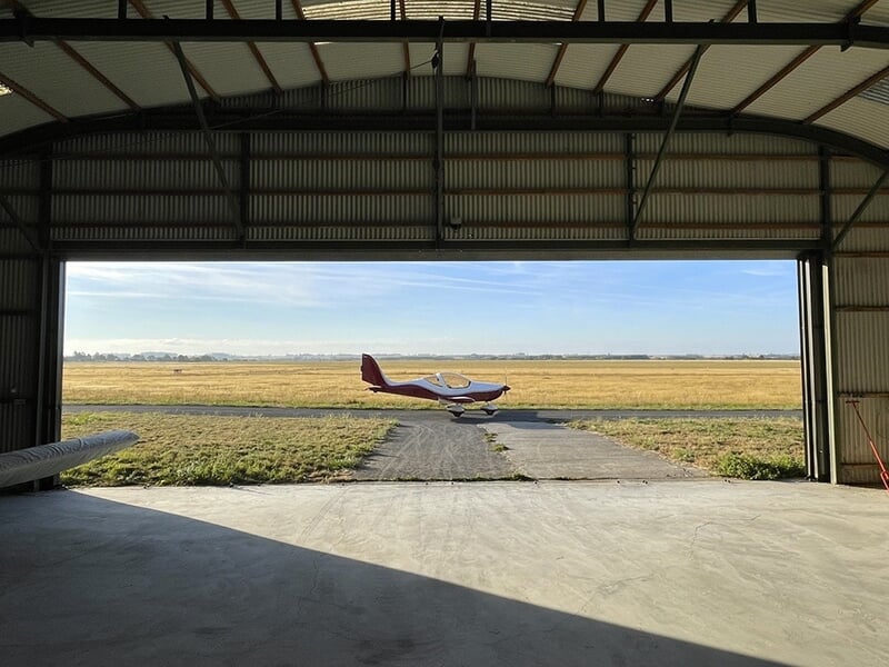 Avion léger visible à travers l'entrée d'un hangar, stationné sur une piste entourée de prairies sous un ciel clair.