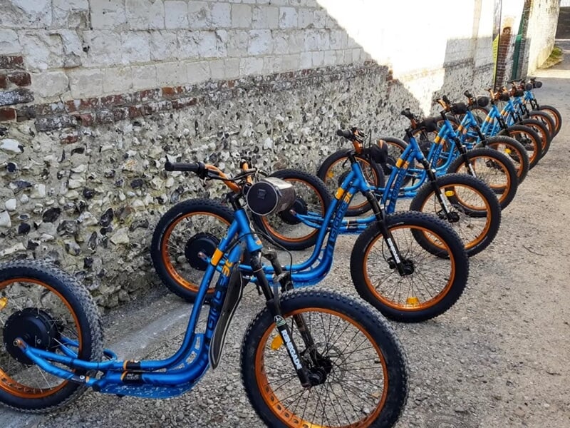 Rangée de trottinettes électriques tout-terrain bleues et orange alignées le long d'un mur en pierre.