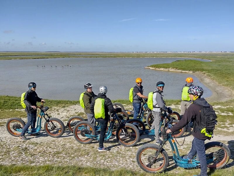 Groupe de personnes posant souriantes avec leurs trottinettes électriques devant un paysage avec de l'eau, des herbes et un ciel dégagé.