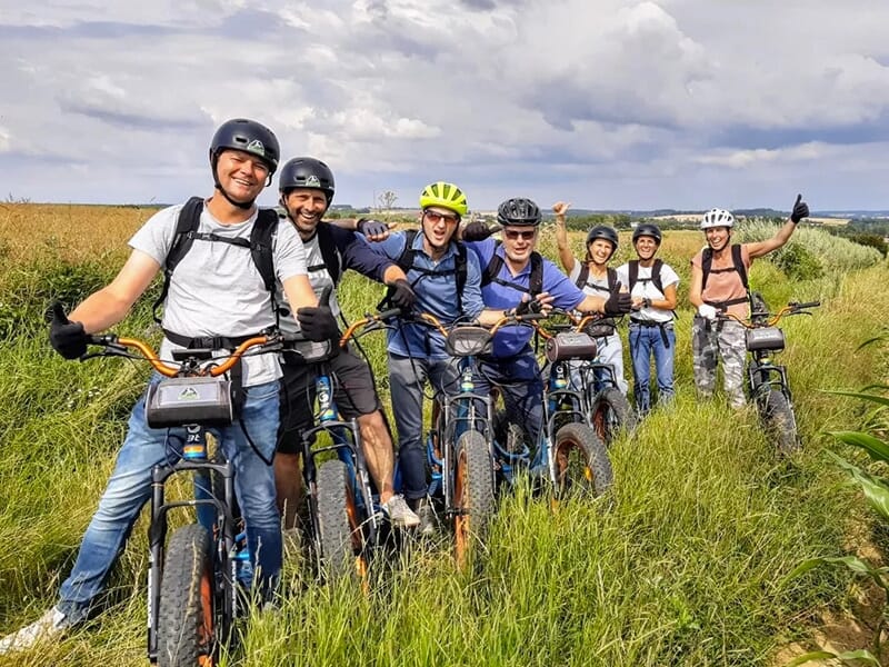 Groupe de personnes souriantes en trottinettes électriques posant sur un chemin bordé de hautes herbes avec un ciel nuageux à l’arrière.