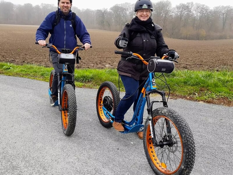 Deux personnes souriantes sur des trottinettes électriques roulant sur une route de campagne près de champs.