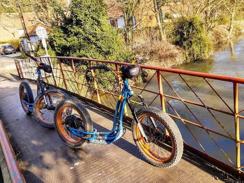 Deux trottinettes électriques stationnées sur un petit pont métallique au-dessus d’une rivière entourée de verdure.