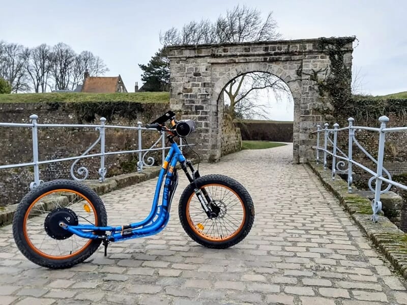 Trottinette électrique tout-terrain stationnée sur un pont pavé en pierre menant à une ancienne arche en pierre, entourée de verdure.