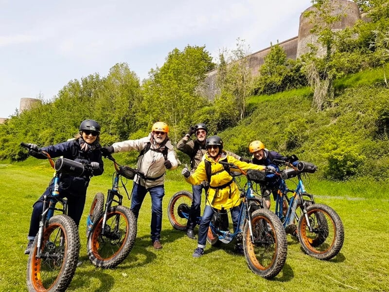Groupe de personnes en trottinettes électriques posant joyeusement devant une pelouse et les remparts d'une fortification historique.