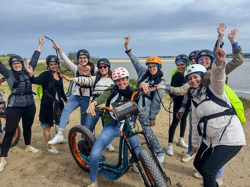 Groupe de femmes souriantes en tenues sportives posant avec enthousiasme autour d’une trottinette électrique sur une plage.