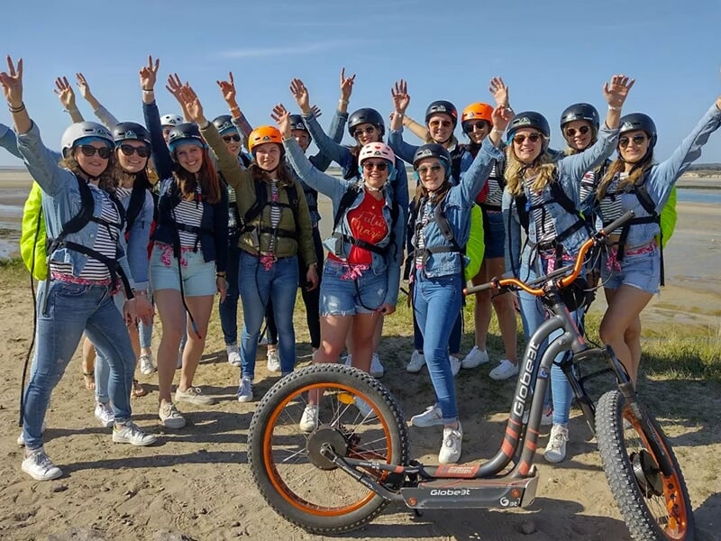 Grand groupe de personnes, majoritairement des femmes, posant joyeusement avec une trottinette électrique sur un rivage sablonneux.