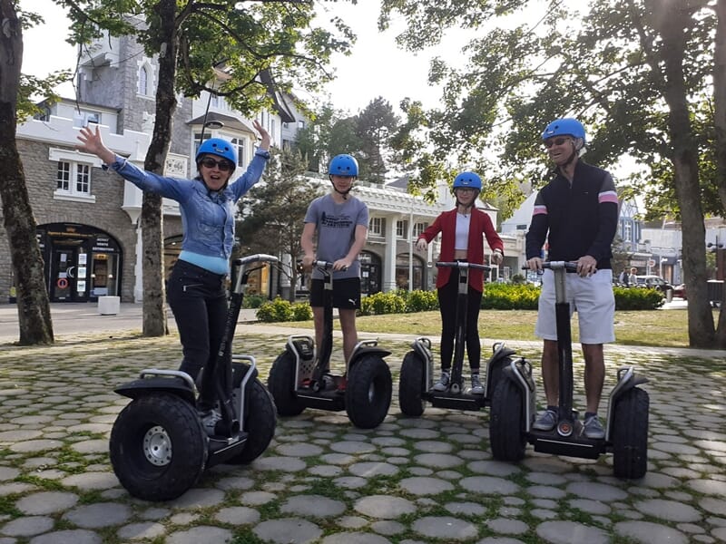 Quatre personnes souriantes portant des casques bleus sur des gyropodes dans un parc avec arbres et bâtiments anciens.