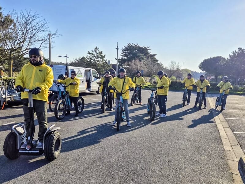 Groupe de personnes en vestes jaunes et casques bleus sur gyropodes roulant sur une route ensoleillée avec arbres.