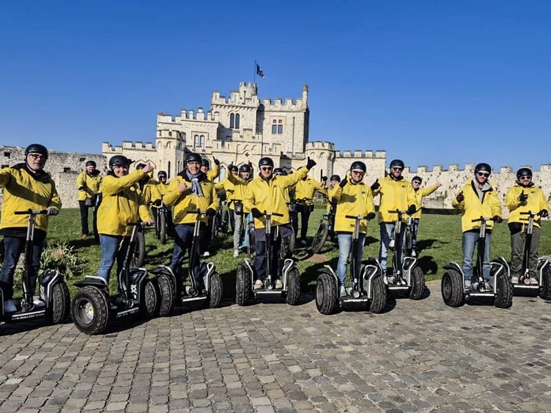 Groupe de personnes en vestes jaunes et casques bleus sur gyropodes posant devant un château sous un ciel bleu.