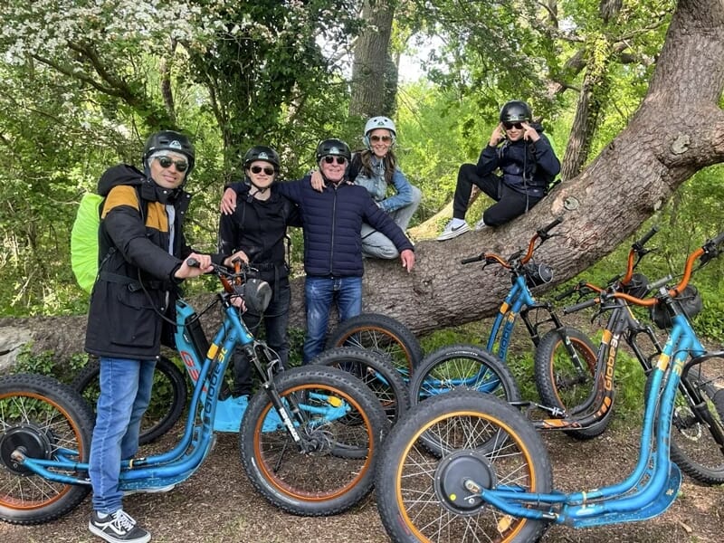 Cinq personnes avec casques posant avec des trottinettes électriques bleues dans un environnement boisé et verdoyant.