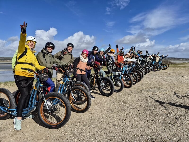Groupe de personnes avec casques sur trottinettes électriques bleues posant sur un terrain sableux sous un ciel nuageux.
