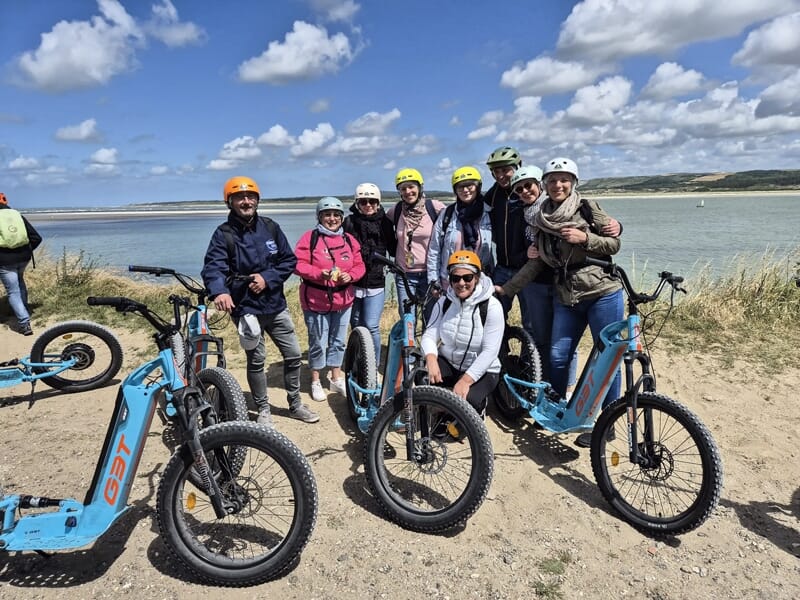 Groupe de personnes souriantes avec casques sur trottinettes électriques bleues sur une plage avec mer et ciel bleu.