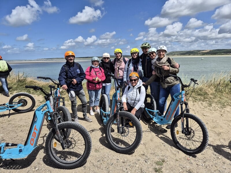 Groupe de personnes souriantes avec casques sur trottinettes électriques bleues sur une plage avec mer et ciel bleu.