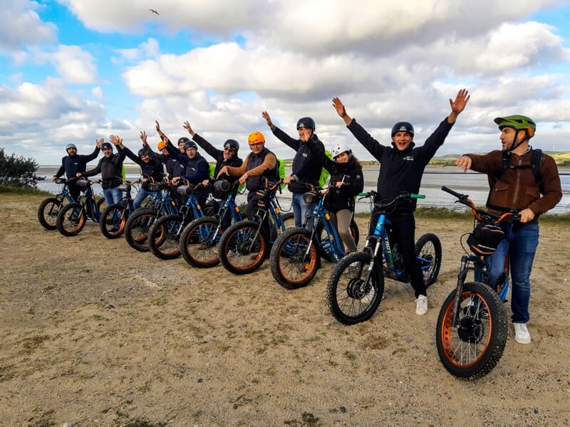 Groupe de personnes avec casques sur trottinettes électriques bleues posant sur un terrain sableux avec ciel partiellement nuageux.