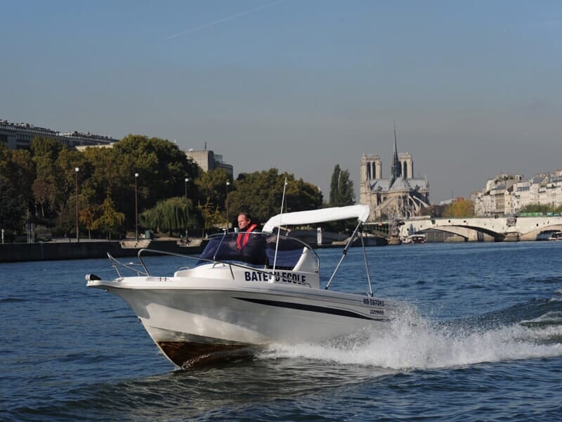 Bateau-école blanc filant sur la Seine avec la cathédrale Notre-Dame de Paris visible en arrière-plan.