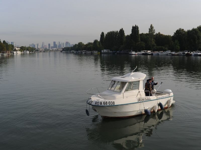 Petit bateau blanc naviguant sur une rivière calme avec des arbres et des bâtiments en arrière-plan sous un ciel clair.