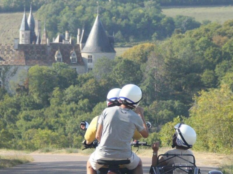 Des motocyclistes sur des motos rétro avec un side-car, roulant sur une route bordée de verdure avec un château en arrière-plan.