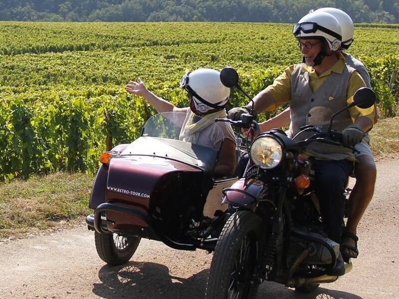 Une moto avec un side-car arrêtée dans une vigne, un conducteur souriant et un passager saluant avec enthousiasme.