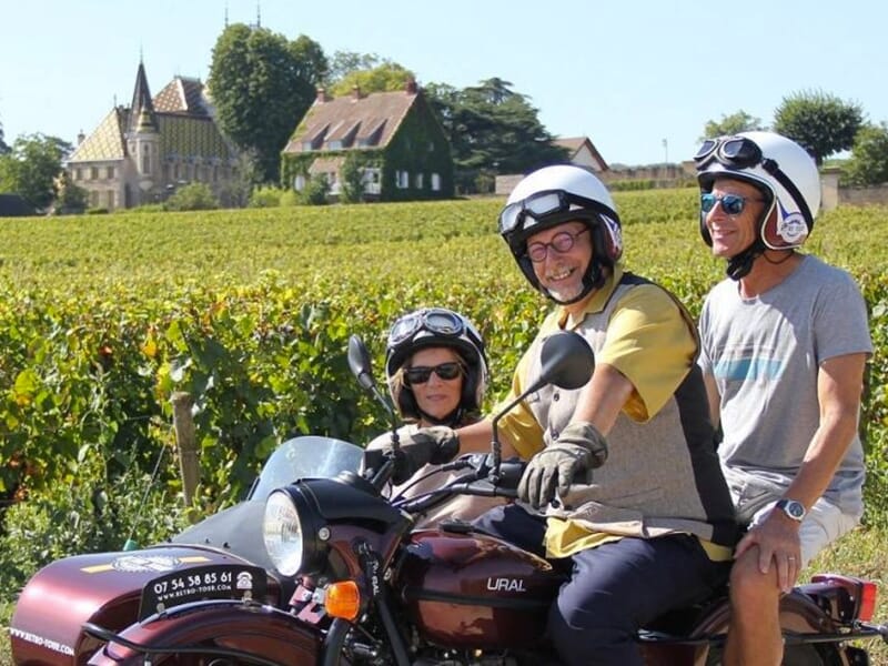 Une moto rétro avec un side-car, devant un paysage de vignes, avec un château en arrière-plan et trois personnes souriantes.