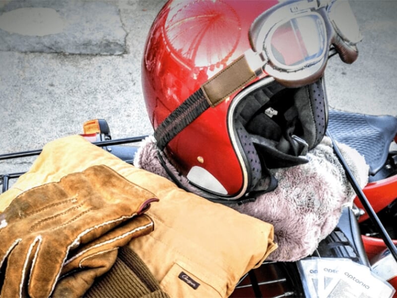 Un casque rouge brillant accompagné de gants en cuir et d’un vêtement posé sur une moto, suggérant une ambiance rétro et un style vintage.