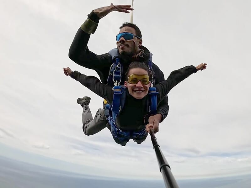 Deux personnes en chute libre en tandem, souriantes, avec un ciel nuageux en arrière-plan.