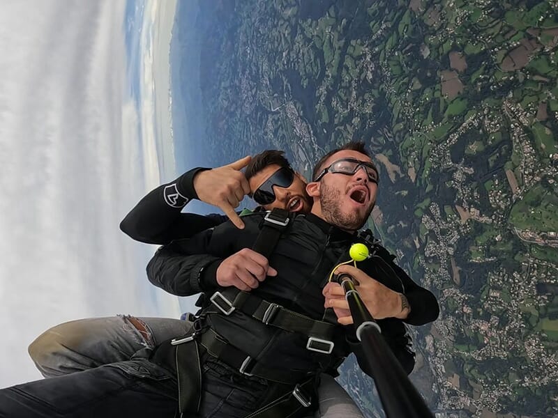 Deux hommes en chute libre en tandem, faisant des gestes joyeux avec la terre visible en dessous.