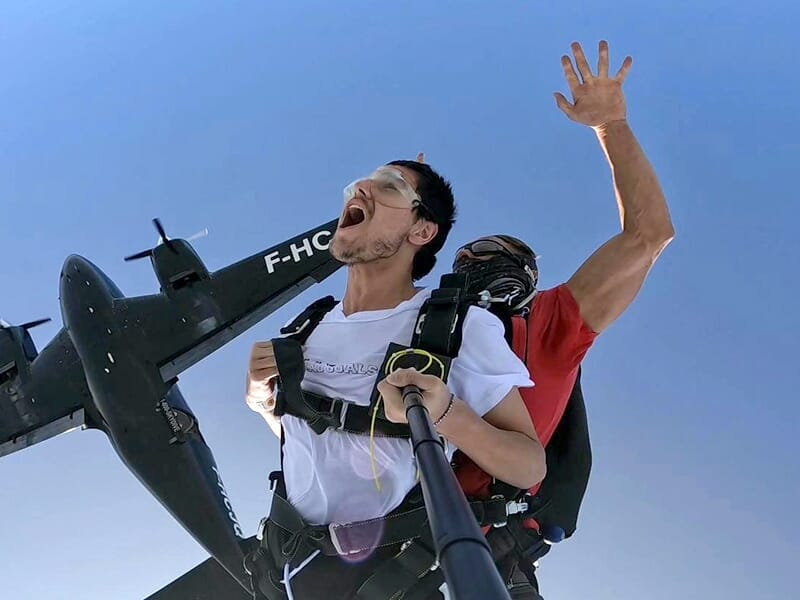 Un tandem de parachutistes criant de joie à la sortie de l’avion, sous un ciel bleu clair.