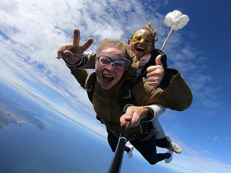 Une femme et son instructeur en plein saut, tous deux souriants et faisant des gestes de victoire au-dessus de l'océan.
