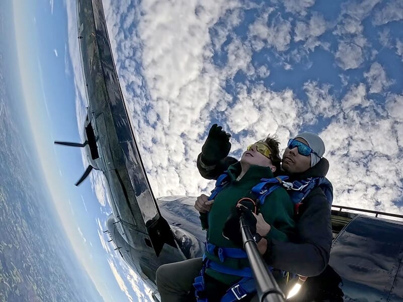 Deux personnes en tandem juste au moment du saut, avec l’avion encore visible à proximité et un ciel partiellement nuageux.