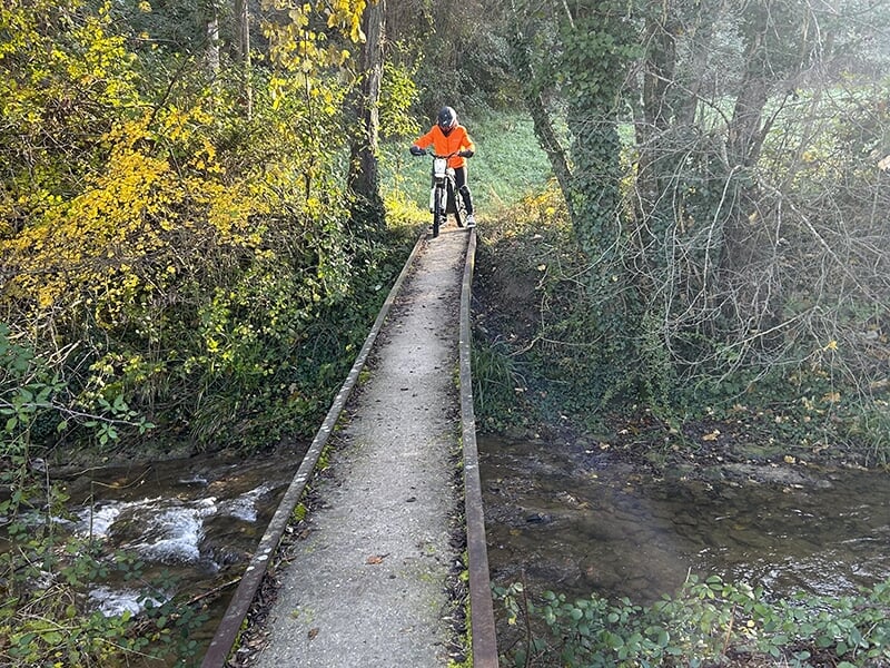 Une personne portant une veste orange traverse un étroit pont en béton avec une trottinette électrique, entouré d'un ruisseau et d'une végétation dense.