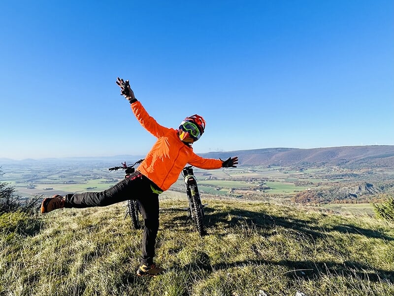 Une personne en veste orange pose joyeusement sur un sommet, à côté d'une trottinette électrique, avec une vue panoramique sur une vallée verdoyante sous un ciel bleu.