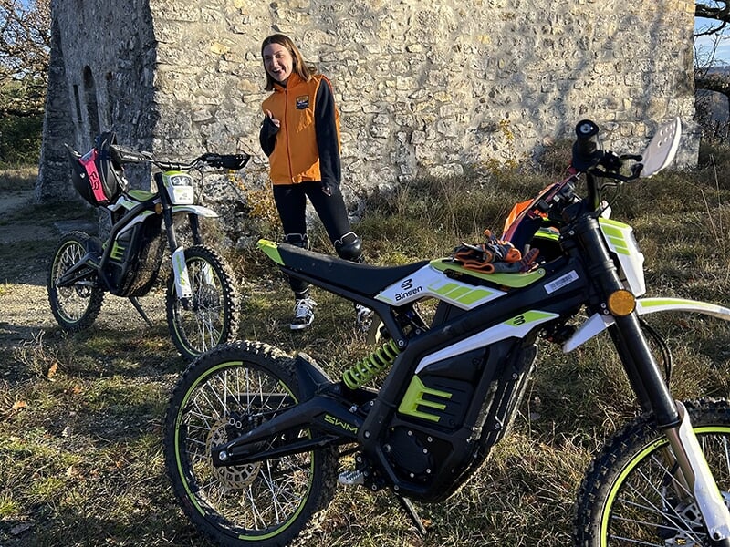 Une femme souriante en veste orange se tient à côté de deux trottinettes électriques, devant un vieux bâtiment en pierre.