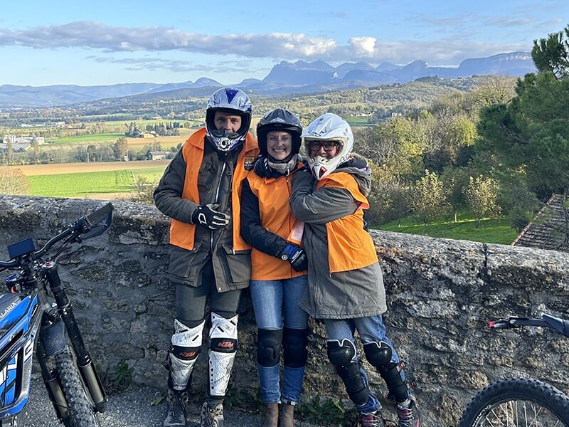 Trois personnes portant des vestes orange et des casques posent ensemble près d'un muret, avec un paysage de campagne et des montagnes en arrière-plan.