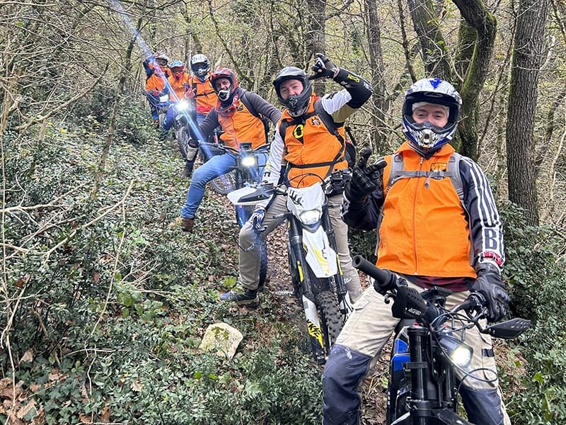 Un groupe de personnes avec des vestes orange et des casques, tenant leurs trottinettes électriques, descend un sentier forestier étroit et verdoyant.