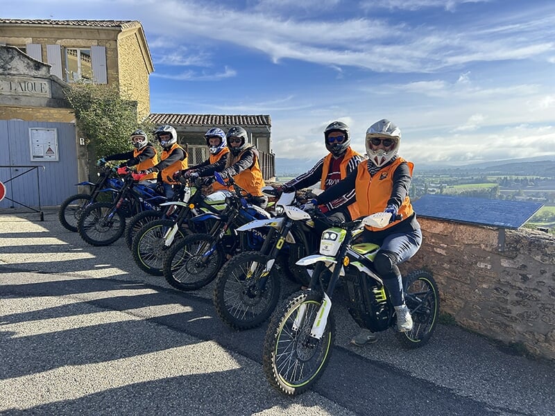 Un groupe de sept personnes en vestes orange et casques pose avec leurs trottinettes électriques devant un bâtiment ancien, sous un ciel dégagé, avec un paysage vallonné à l'horizon.