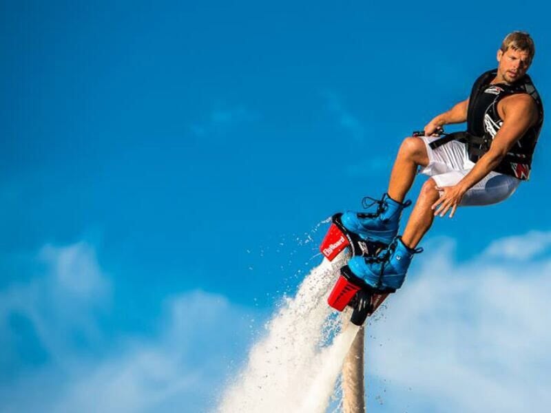 Homme en short blanc et gilet de sauvetage volant au-dessus de l'eau avec un flyboard sous un ciel bleu.