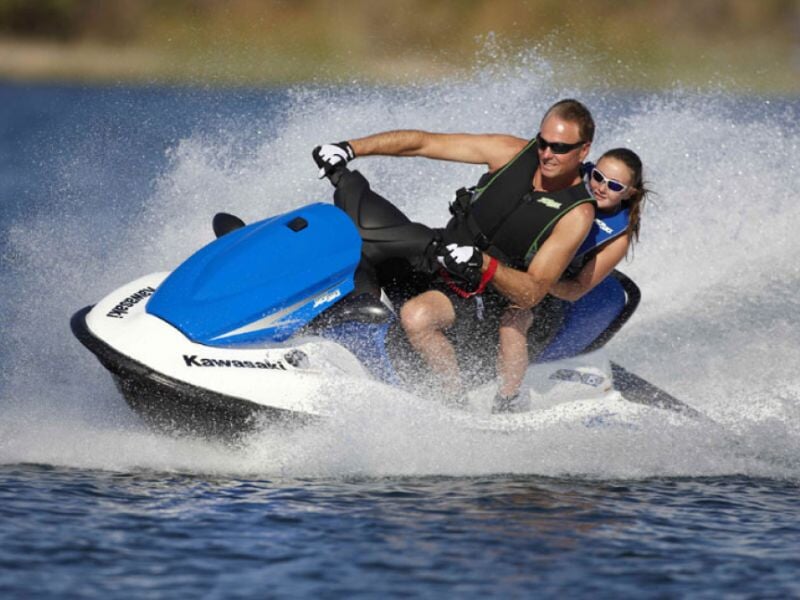 Homme et femme en gilets de sauvetage sur un jetski Kawasaki bleu et blanc avec éclaboussures d'eau.
