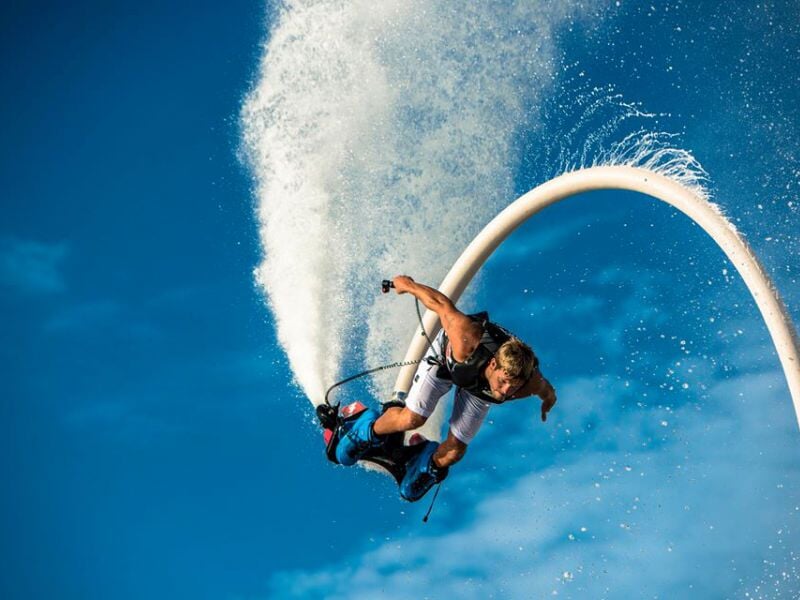 Homme en short et gilet de sauvetage utilisant un flyboard propulsé par de l'eau sous un ciel bleu.